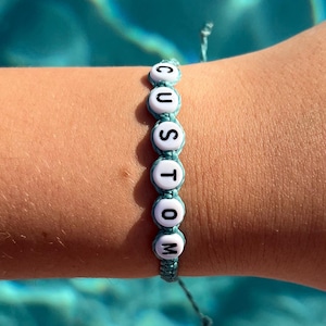 May include: A turquoise blue friendship bracelet with white beads that spell out the word "CUSTOM".