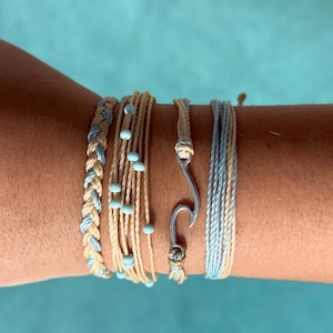 May include: A set of five bracelets in shades of blue and beige. The bracelets are made of string and beads. One bracelet features a silver hook charm.