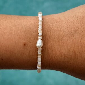 May include: A white pearl bracelet with a single large pearl charm. The bracelet is made of small white and beige beads.