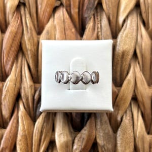 May include: A silver ring featuring a repeating seashell design. The ring is displayed on a white surface against a woven, textured background. The ring's design is detailed and the metal has a polished finish.