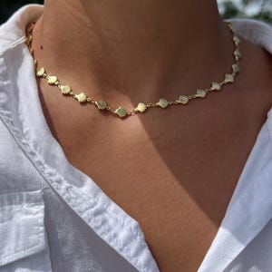 May include: A gold-colored necklace featuring a repeating seashell design. The necklace is worn against a white collared shirt. The seashells are evenly spaced and connected by small links, creating a delicate and elegant look.