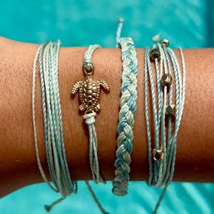 May include: A collection of five bracelets in shades of blue and white. One bracelet features a gold-colored turtle charm. Another is a braided design, and a third has gold-colored beads. The bracelets are tied with adjustable knots.