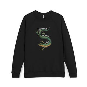 May include: Black long-sleeve sweatshirt featuring a green and yellow newt illustration with the text "She turned me into a Newt." The design is centered on the chest. The sweatshirt has a crew neck and raglan sleeves.