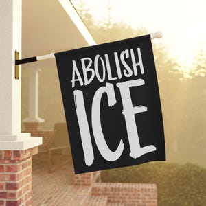 May include: Black flag with the words "ABOLISH ICE" in white, bold, handwritten-style font. The flag is mounted on a white pole and displayed outside a building with brick and white accents.