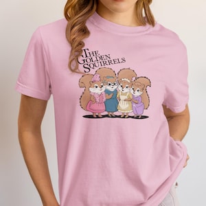 May include: A pink t-shirt with a graphic of four cartoon squirrels wearing dresses. The text "The Golden Squirrels" is above the squirrels.