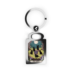 May include: Silver-tone keychain with a rectangular charm featuring a whimsical illustration. The artwork depicts black cats gathered around a cauldron in a forest setting under a full moon. The text "THE COVEN" is at the bottom.
