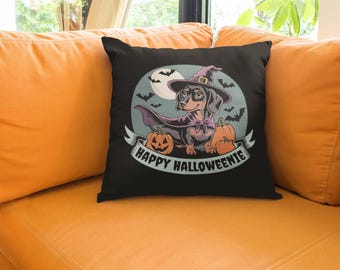 May include: Black decorative throw pillow featuring a dachshund dressed as a witch for Halloween. The design includes a witch hat, cape, pumpkins, bats, and the text "Happy Halloweenie" on a banner.