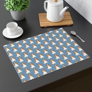 May include: A rectangular placemat with a repeating pattern of Cavalier King Charles Spaniel illustrations on a light blue background. The dogs are depicted in shades of brown, white, and tan. The placemat is on a dark surface.