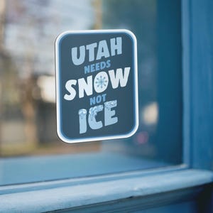 May include: A rectangular sticker with rounded corners, featuring the text "UTAH NEEDS SNOW NOT ICE" in white and light blue. The sticker has a dark blue background and a light blue border, with a snowflake graphic in the word "SNOW".