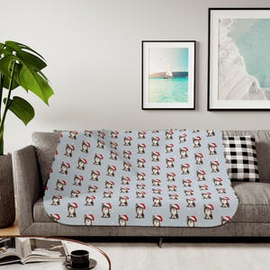 May include: A light blue blanket with a repeating pattern of dogs wearing Santa hats and snowflakes. The blanket is draped over a gray sofa, with framed artwork and a decorative pillow in the background.