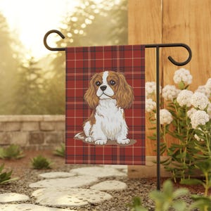 May include: A garden flag featuring a Cavalier King Charles Spaniel illustration. The dog is depicted with brown and white fur, sitting against a red plaid background. The flag is displayed on a black metal stand, surrounded by greenery.