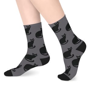 May include: A pair of mid-calf socks in a dark gray color featuring a repeating pattern of black cat silhouettes. Each cat has white eyes and is depicted in a sitting position. The socks have solid black toe and heel sections for contrast.