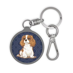 May include: A silver-toned keychain with a round charm featuring a Cavalier King Charles Spaniel illustration. The dog has brown and white fur, sitting on a brown surface against a dark blue patterned background. The keychain has a clasp and a key ring.