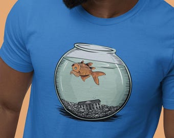 May include: A bright blue t-shirt featuring a graphic illustration of a round glass fishbowl filled with water, containing a single orange goldfish swimming near the top and a small architectural building structure resting on the gravel base.