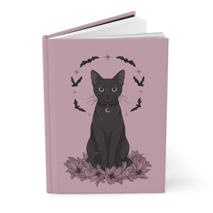 May include: A hardcover journal with a dusty rose cover featuring a centered illustration of a black cat wearing a crescent moon pendant. The cat is surrounded by a circular arrangement of small black bats and stars, with a cluster of pink flowers at its base.