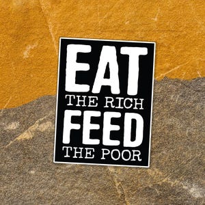 May include: A black and white sticker with the text "EAT THE RICH FEED THE POOR". The sticker has a rectangular shape with rounded corners and is placed on a textured background of brown and gray.