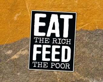 May include: A black and white sticker with the text "EAT THE RICH FEED THE POOR". The sticker has a rectangular shape with rounded corners and is placed on a textured background of brown and gray.