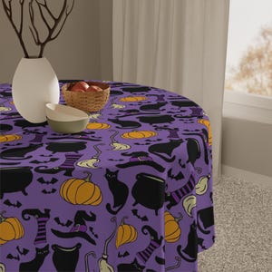 May include: A round purple Halloween-themed tablecloth featuring a repeating pattern of black cats, pumpkins, witch hats, cauldrons, and bats. The tablecloth is on a round table with a white vase, a bowl, and a basket of fruit.