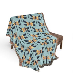 May include: A light blue throw blanket featuring a repeating pattern of dragonflies in shades of yellow, orange, and navy blue. The blanket has a fringed edge and is draped over a bench.