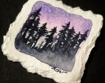 May include: A small watercolor painting of a forest silhouette against a purple and pink sky. The artwork is on a square piece of textured white paper with a deckled edge. The artist's signature is in the lower right corner.