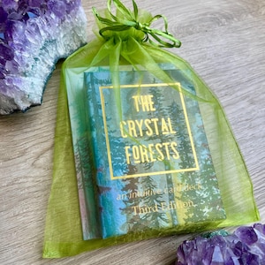 以下が含まれることがあります： 「The Crystal Forests」の直感的なカードのデッキ。緑色のオーガンジーバッグに入っており、金色の文字と森の風景が描かれています。アメジストの結晶がバッグの両側に表示されています。第三版が確認できます。