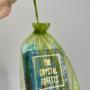 May include: A green organza gift bag contains a card deck labelled "The Crystal Forests." The card deck features a forest scene design with gold lettering. The bag is held by a green stem.