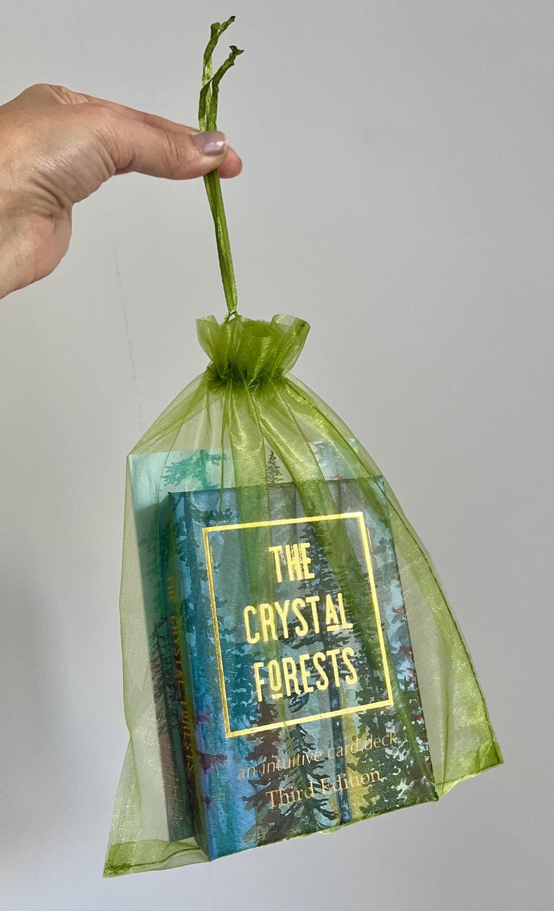 May include: A green organza gift bag contains a card deck labelled "The Crystal Forests." The card deck features a forest scene design with gold lettering. The bag is held by a green stem.