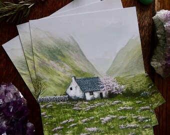 May include: A stack of landscape art prints featuring a white cottage with a green roof nestled in a valley. The prints depict a field of purple flowers, green hills, and a cloudy sky. The prints are on a wooden surface.