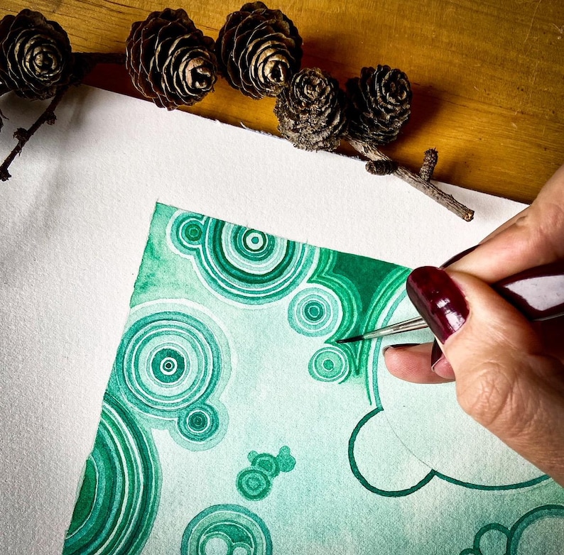 May include: A watercolour painting in shades of green featuring concentric circles and organic shapes. A hand with a paintbrush is adding details to the artwork. Pine cones and a branch are arranged on a wooden surface.