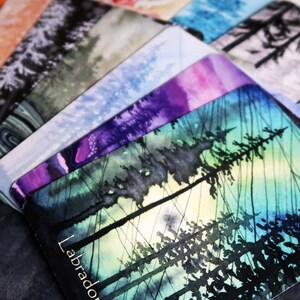 May include: A selection of watercolour art cards depicting forest scenes in diverse colours. The cards showcase a spectrum of hues, including blues, purples, and greens, with the word "Labradorite" visible on one card.