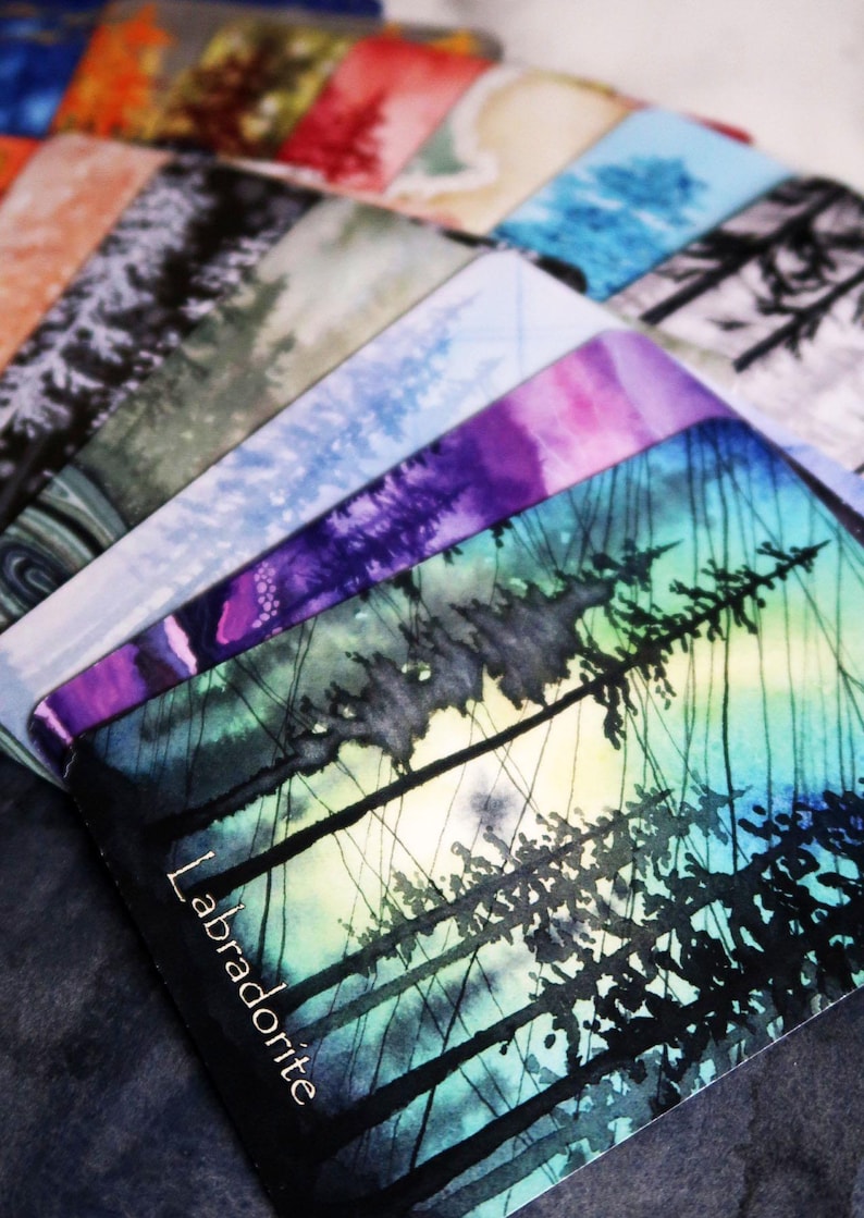 May include: A selection of watercolour art cards depicting forest scenes in diverse colours. The cards showcase a spectrum of hues, including blues, purples, and greens, with the word "Labradorite" visible on one card.