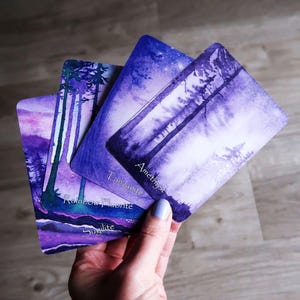 May include: Three purple and blue oracle cards with watercolour forest scenes. The cards are labelled "Rainbow Fluorite", "Sugilite", and "Amethyst", and "Tanzanite". The cards are held in a hand with light blue nail polish.