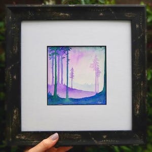 May include: A framed watercolour painting depicting a forest scene. The artwork shows tall, slender trees in shades of purple and green against a soft, gradient sky. The frame is black with a distressed finish.