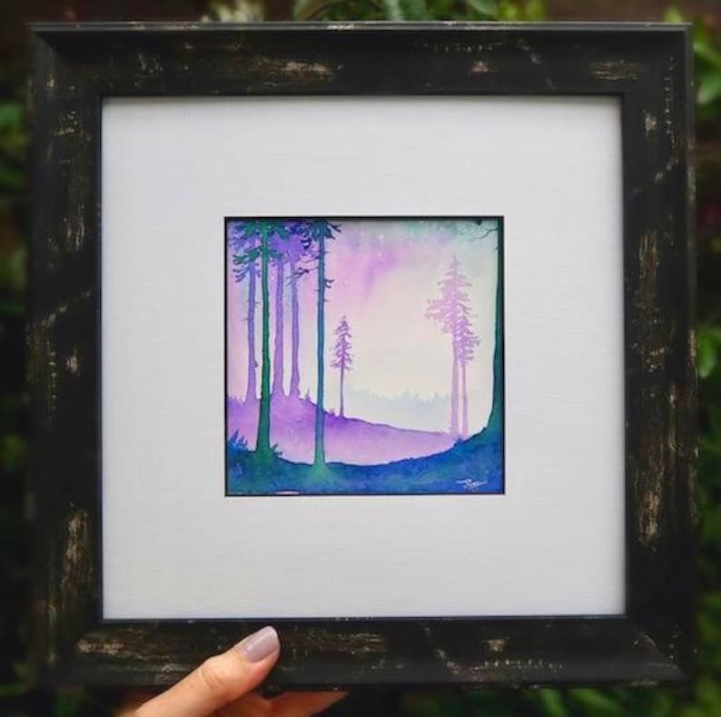 May include: A framed watercolour painting depicting a forest scene. The artwork shows tall, slender trees in shades of purple and green against a soft, gradient sky. The frame is black with a distressed finish.