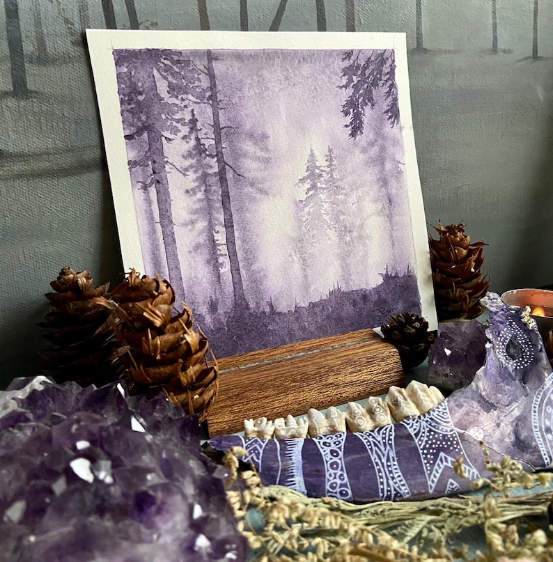 May include: A watercolour painting of a misty forest in shades of purple and grey. The artwork is displayed with pine cones, amethyst crystals, and a decorated jawbone. The scene evokes a mystical, natural theme.