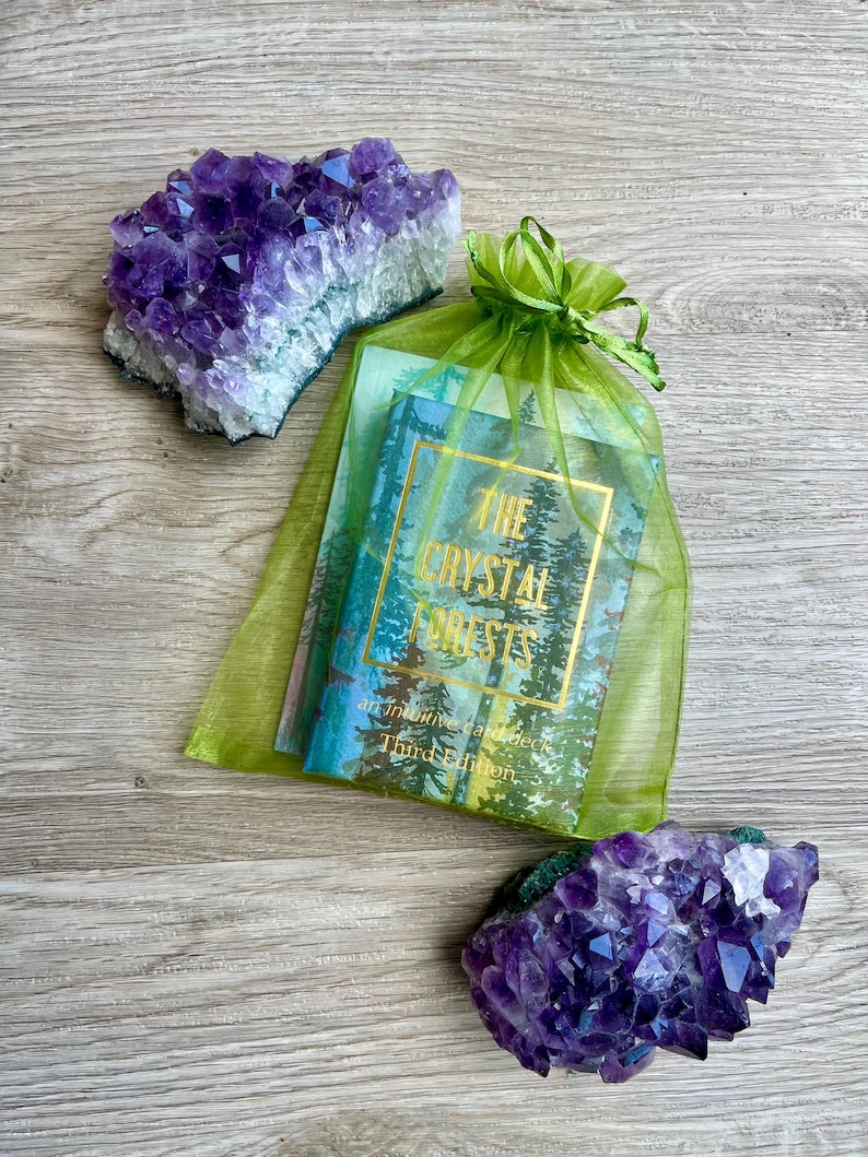 May include: Two amethyst crystal clusters and a deck of cards in a green organza bag. The card deck has a forest scene and the text "The Crystal Forests" in gold lettering. The crystals are a deep purple colour.