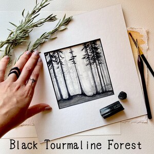May include: A monochrome watercolour painting of a forest scene, framed in white. The painting is displayed with a sprig of rosemary, a black ring, two paintbrushes, and two pieces of black tourmaline. The text "Black Tourmaline Forest" is at the bottom.