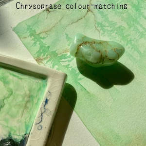 May include: A pale green chrysoprase gemstone with brown markings rests on a watercolour painting. The painting displays green and brown tones, with the text "Chrysoprase colour-matching" at the top. A white ceramic tray with green and blue paint is in the lower left corner.
