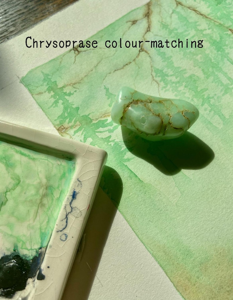 May include: A pale green chrysoprase gemstone with brown markings rests on a watercolour painting. The painting displays green and brown tones, with the text "Chrysoprase colour-matching" at the top. A white ceramic tray with green and blue paint is in the lower left corner.