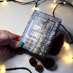 May include: A deck of cards titled "The Crystal Forests" with a forest scene design. The box is held by a hand with a teal painted nail. Pine cones and string lights are scattered around the box.