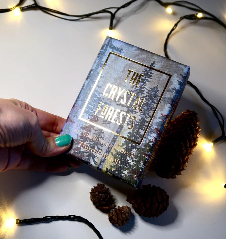 May include: A deck of cards titled "The Crystal Forests" with a forest scene design. The box is held by a hand with a teal painted nail. Pine cones and string lights are scattered around the box.