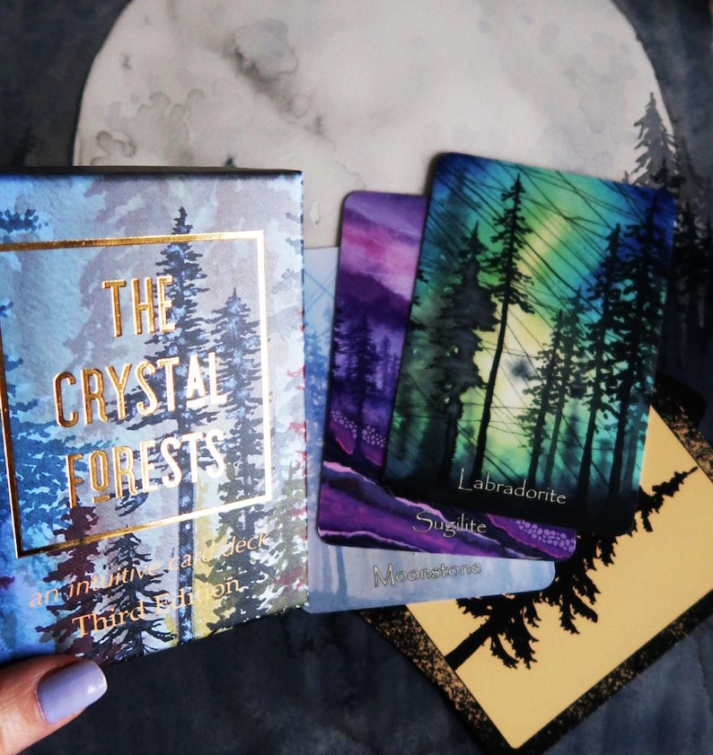 May include: An oracle card deck titled "The Crystal Forests". The box and cards display forest imagery with gold lettering. Three cards are visible, labelled "Labradorite", "Sugilite", and "Moonstone". The colour scheme includes blues, greens, and purples.