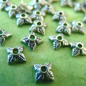 May include: Silver-coloured metal leaf-shaped beads with a central hole. The beads have a four-leaf clover design with detailed leaf veins. The beads are scattered on a green surface, creating a textured appearance.