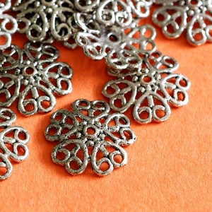 May include: Close-up of several silver filigree flower-shaped components. Each component has a detailed, openwork design with a central circle and six curved petals. The components are scattered on an orange surface.