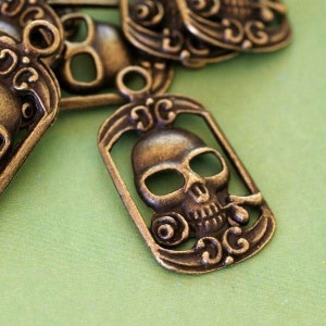 May include: Antique brass-coloured skull pendants with decorative frames and loops for jewellery making. The pendants feature detailed skull designs with open eye sockets and ornate scrollwork. The background is a light green fabric.