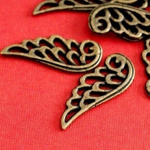 May include: Close-up of several antique bronze-coloured metal wing charms. Each wing has a detailed, openwork design with curved lines and swirls. The charms are arranged on a red textured surface.