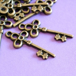May include: Close-up of several antique bronze-coloured keys with a flower-shaped top and a decorative key bit. The keys are scattered on a light purple surface. The keys are likely for crafting or jewellery making.