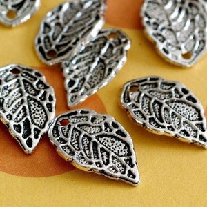 May include: Close-up of several silver leaf charms. Each charm has detailed veining and a small hole at the top for stringing. The charms are scattered on a yellow surface, creating a natural, botanical theme.