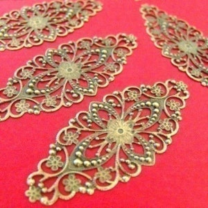 May include: Ornate, filigree metal embellishments in a bronze tone, arranged on a red background. Each piece features a central floral design with surrounding delicate, openwork patterns and small beaded accents. The pieces are likely for crafting or jewelry making.