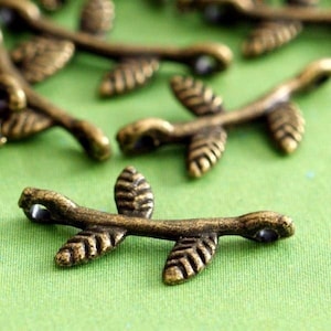 May include: Close-up of several bronze-coloured leaf charms. Each charm features a detailed leaf design with a small hole at each end. The charms are arranged on a green surface, suggesting a craft or jewellery-making theme.